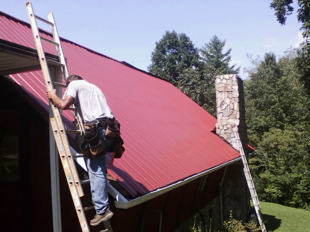Roofer on a ladder beside a red metal roof and stone chimney — Soffit & Fascia Repair by Roofing Brothers in Mongaup Valley
